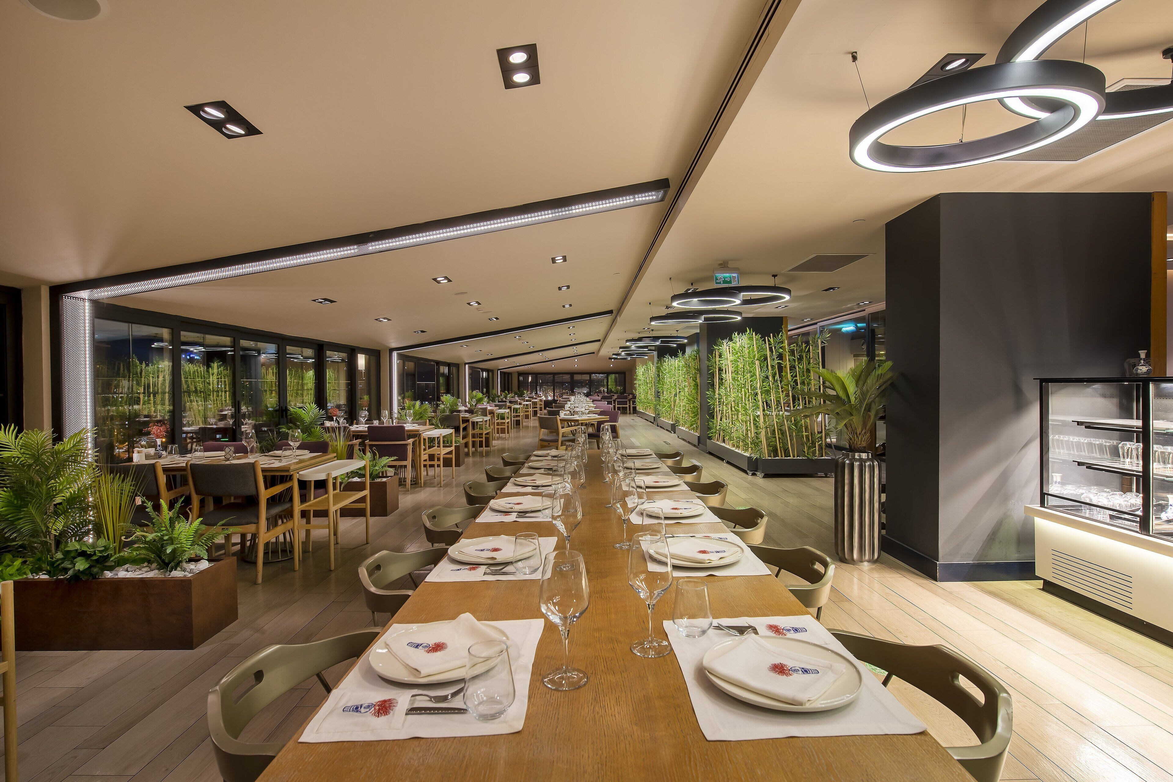 A modern restaurant with long tables set for dining, surrounded by indoor plants and large windows.