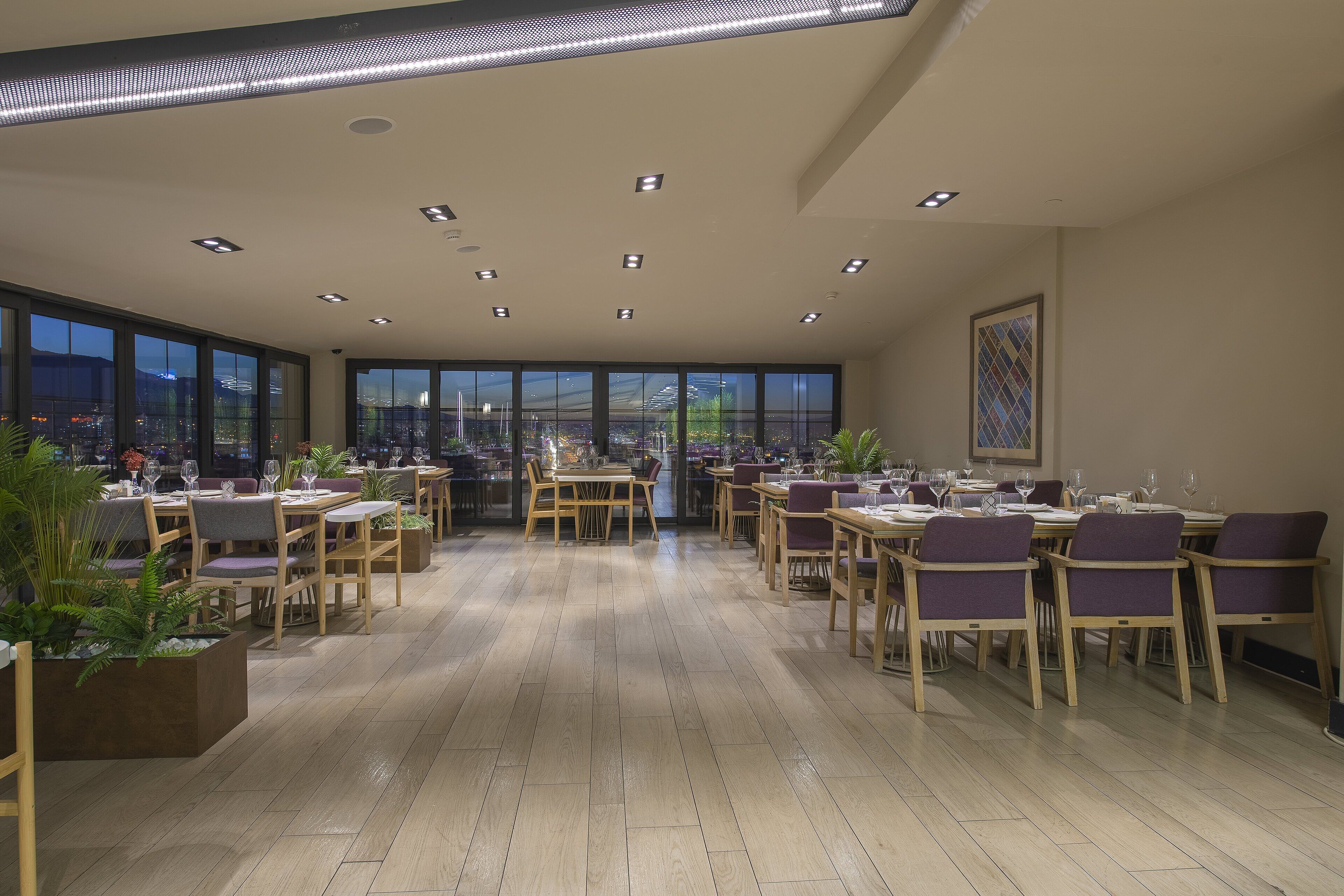 A spacious dining area with multiple tables and chairs, large windows offering a city view, and recessed ceiling lighting.