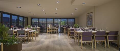A spacious dining area with multiple tables and chairs, large windows offering a city view, and recessed ceiling lighting.