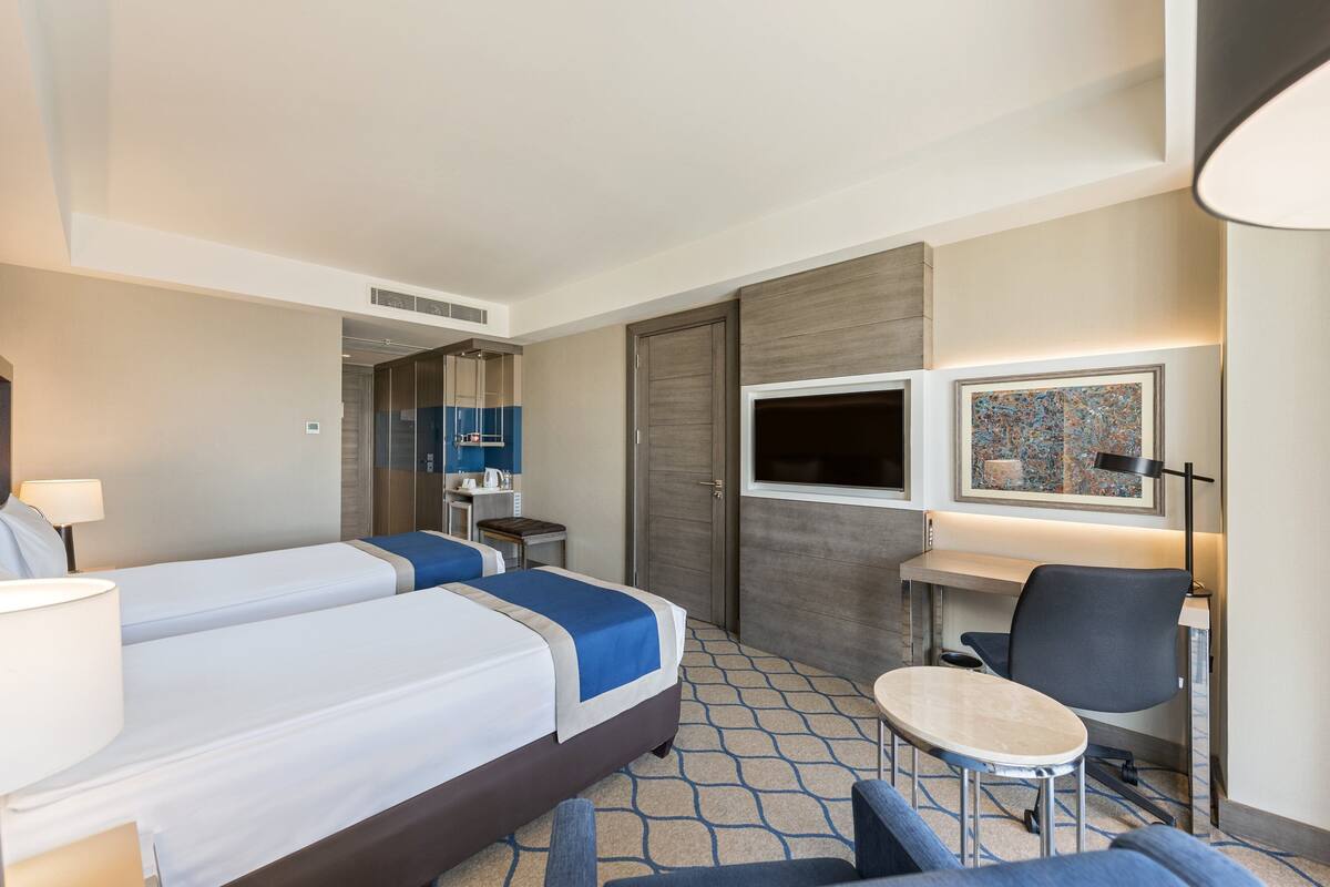 A hotel room with two beds, a desk, a chair, a small table, a lamp, a TV, and a painting on the wall.