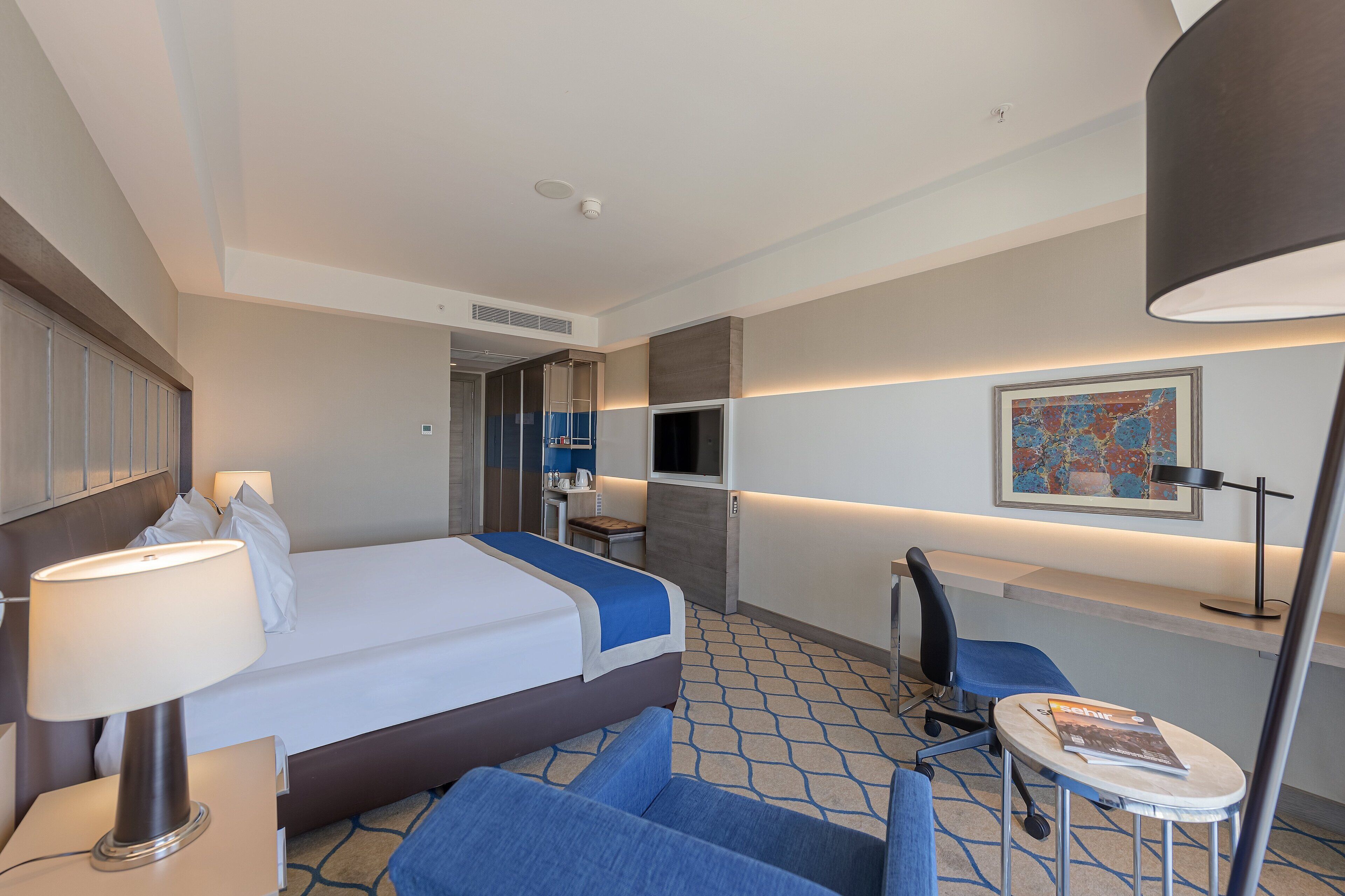 A hotel room with a bed, a desk, a chair, a lamp, and a TV.