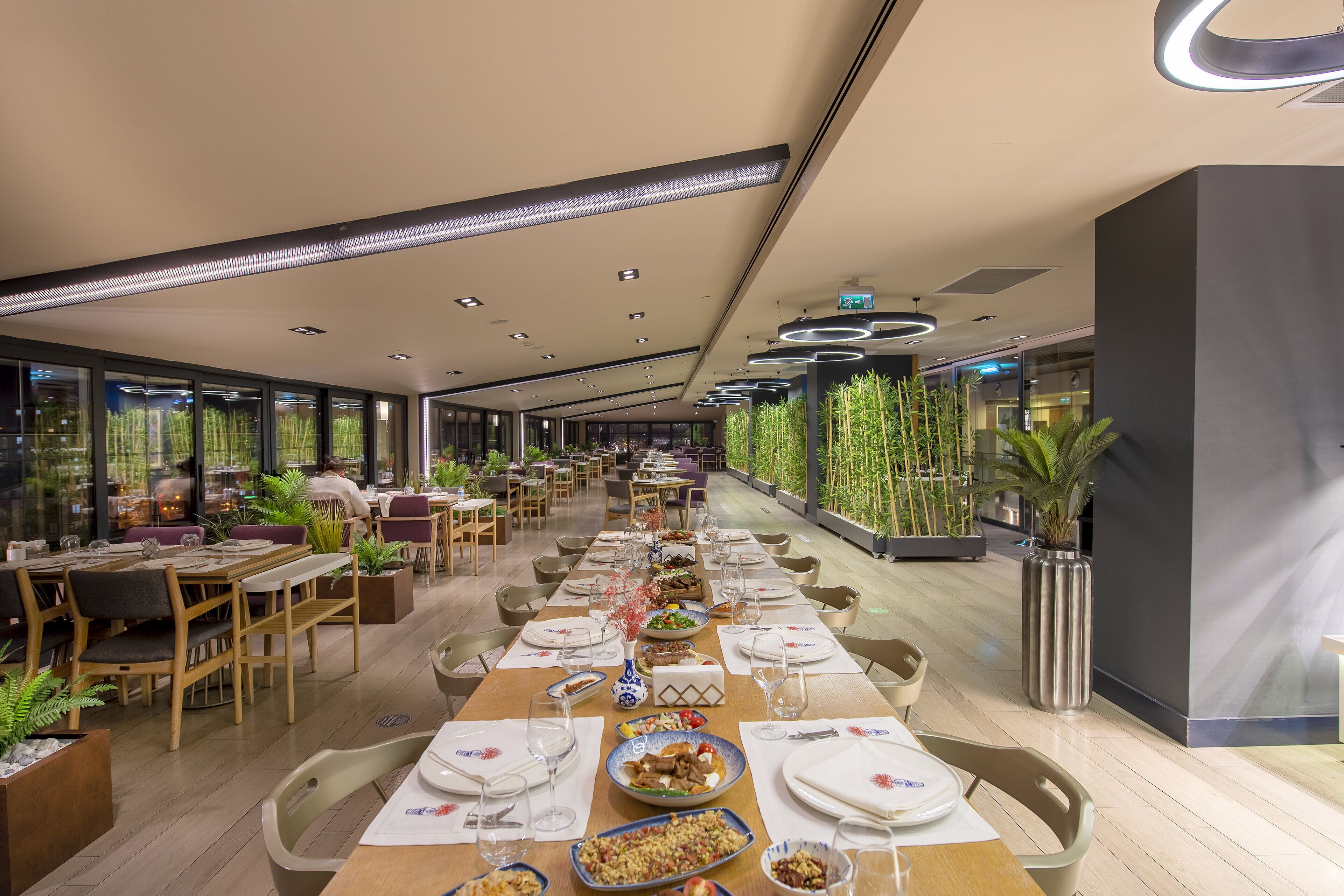A modern restaurant with long tables, wooden chairs, and potted plants.