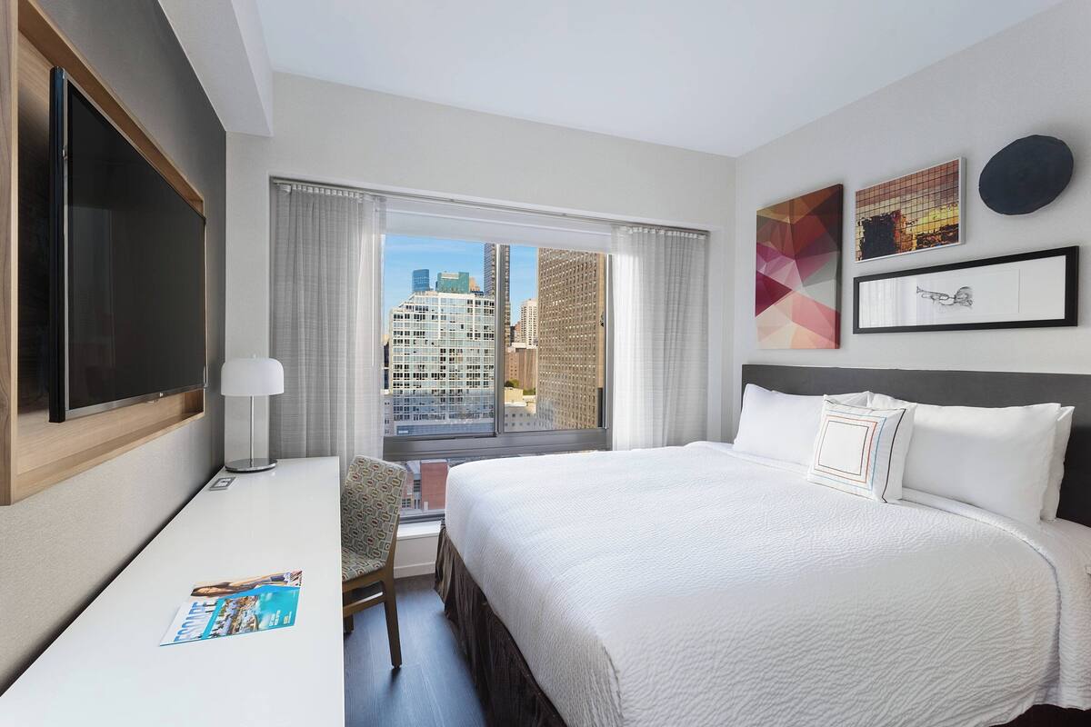 Une chambre d’hôtel équipée d’un lit, d’un bureau, d’une chaise, d’une télévision et d’une fenêtre donnant sur la ville.