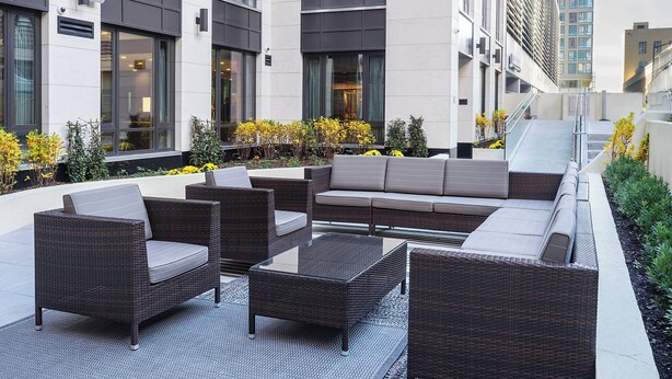 Un área de estar al aire libre con muebles de mimbre y una mesa con superficie de vidrio, rodeada por un edificio con grandes ventanales y una escalera.