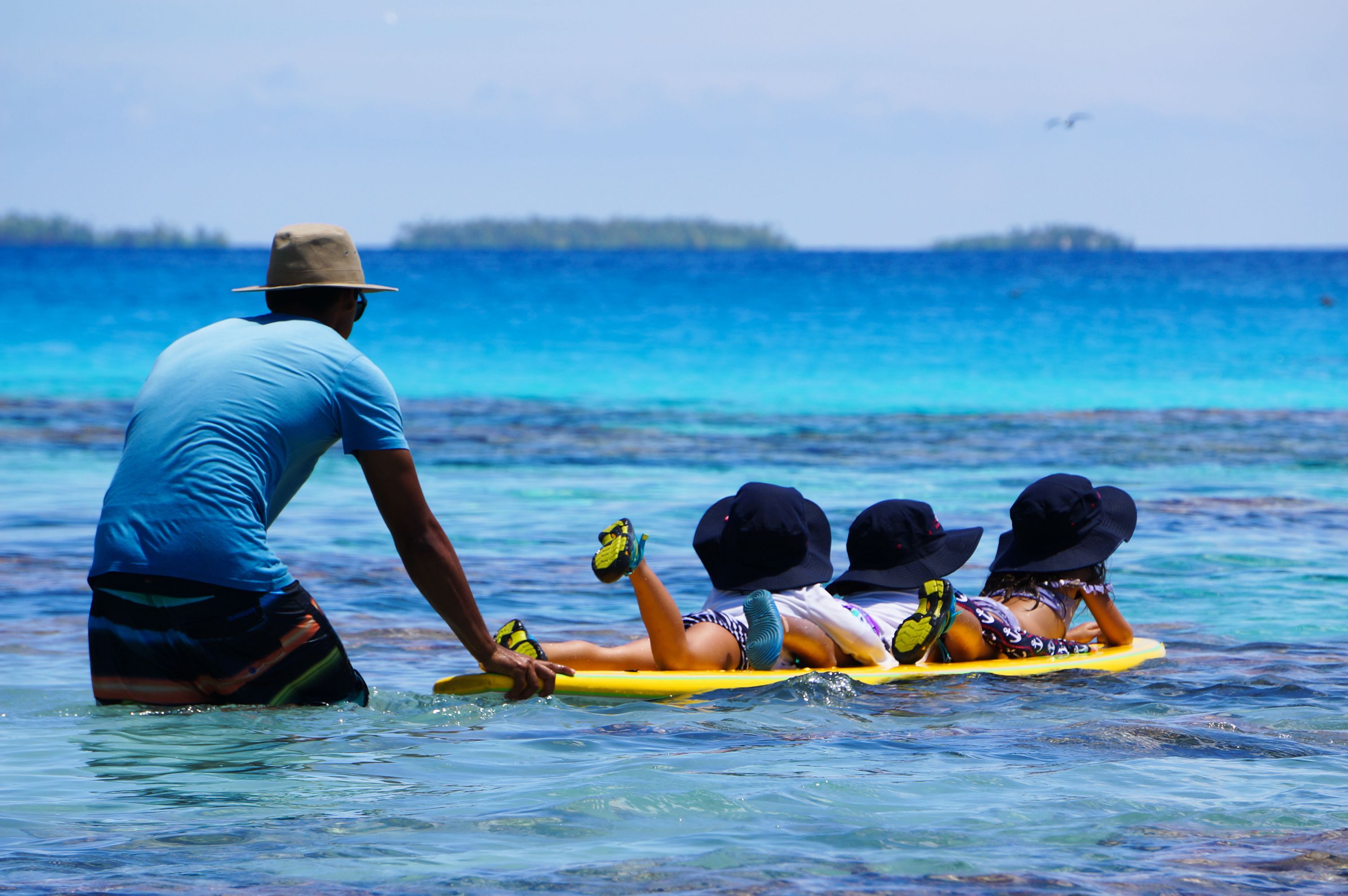 Una persona con camisa y gorra azul ayuda a dos personas sobre una tabla de surf amarilla en el océano.