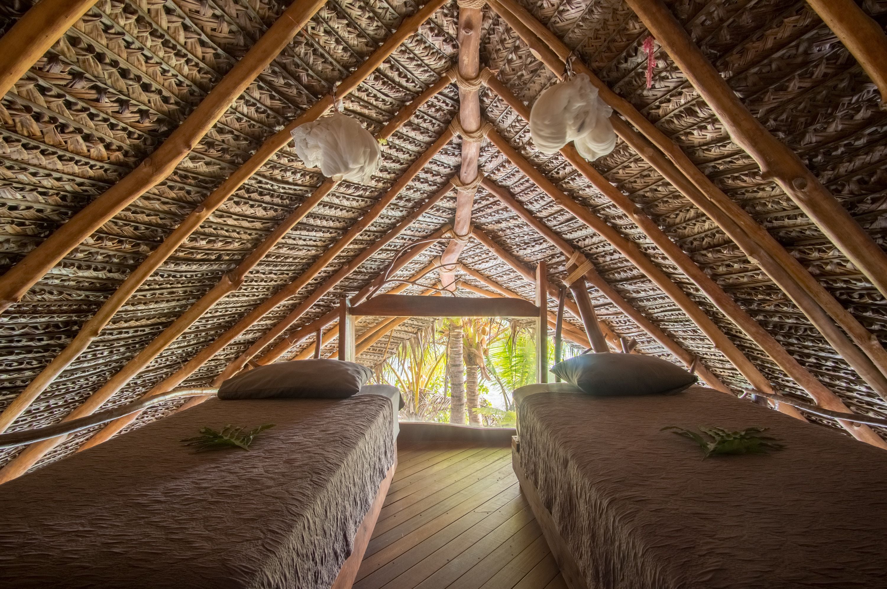 A thatched-roof room with two beds, wooden beams, and a view of palm trees.