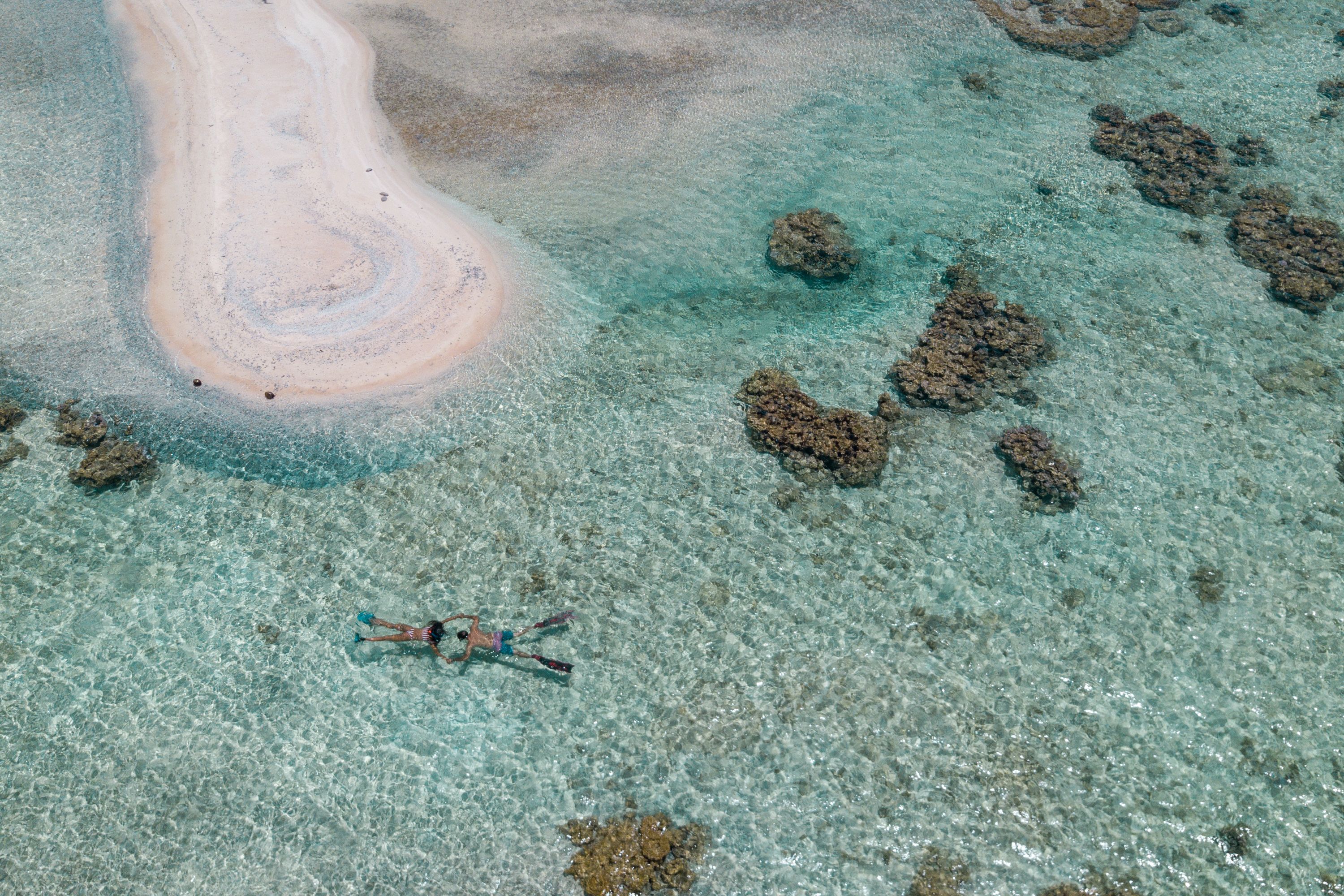 Vue aérienne d’une plage avec une eau turquoise et limpide, une zone sablonneuse et quelques rochers épars.