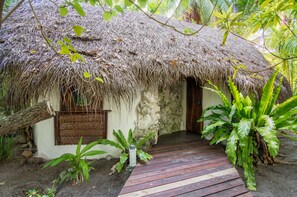 A thatched-roof hut with a wooden door and a small window, surrounded by lush greenery. - Ninamu Resort (Tikehau)