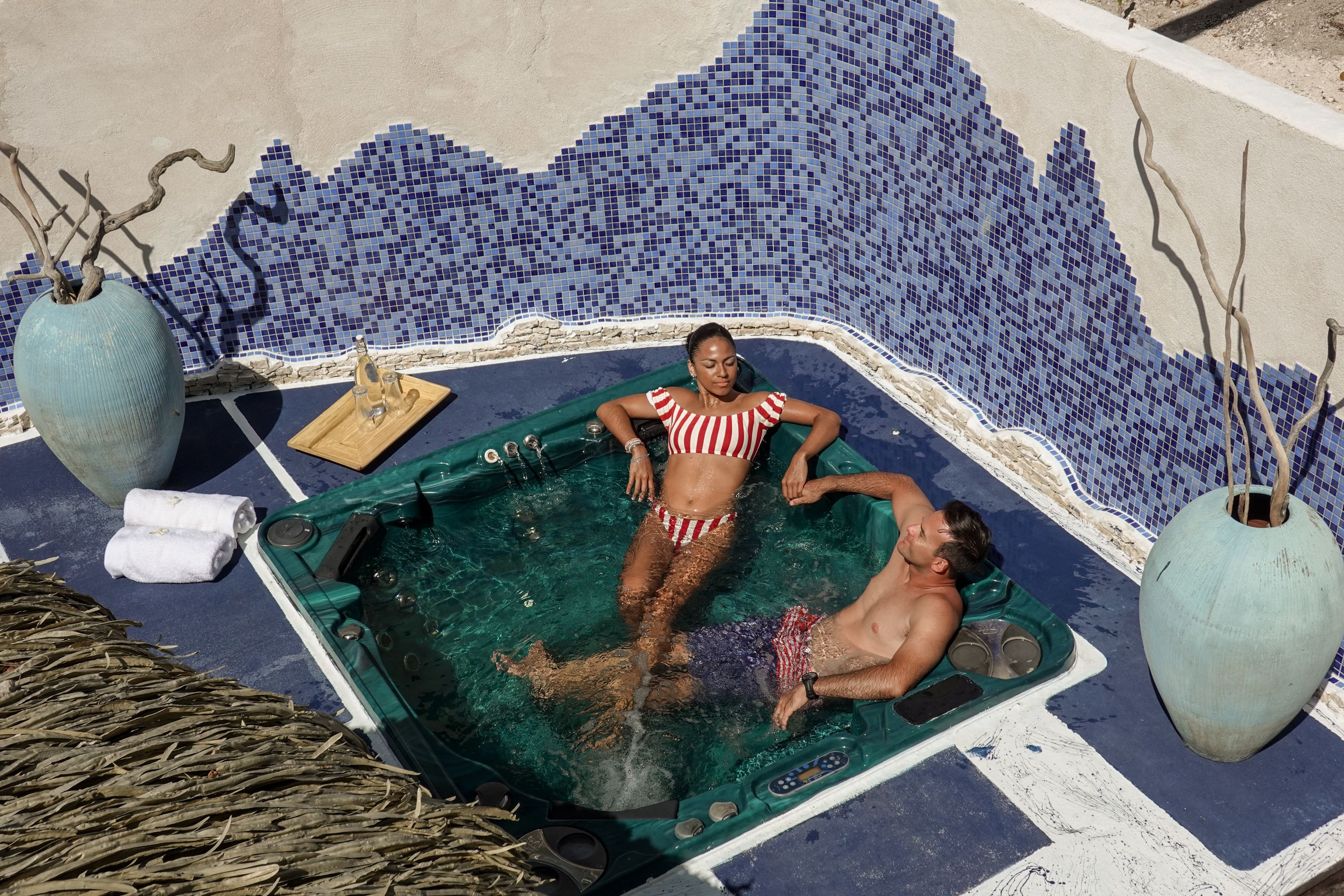 A hot tub with mosaic tile walls, two large vases, and a wooden tray with a glass and a towel.