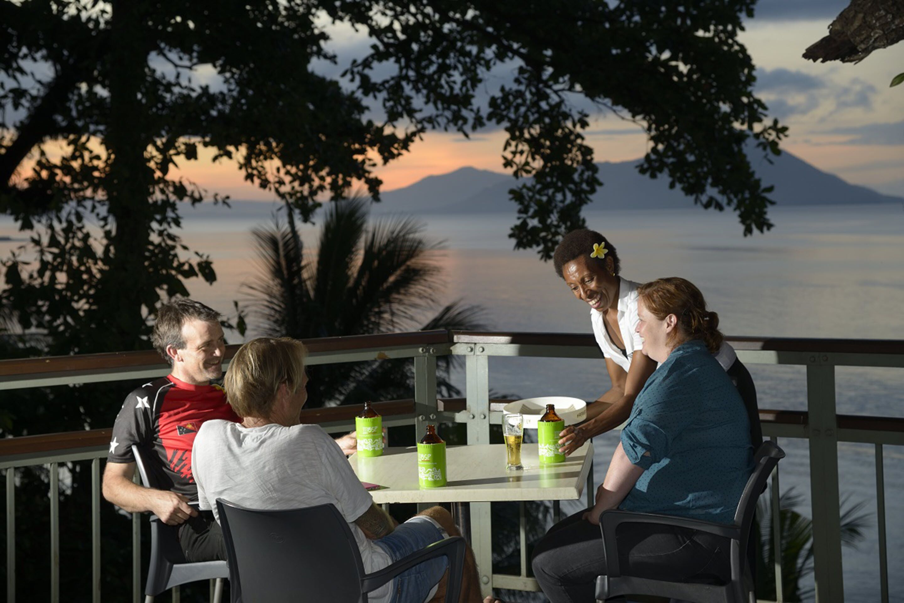 Un grupo de personas sentadas en una mesa al aire libre, con una vista panorámica de un cuerpo de agua y montañas al fondo.