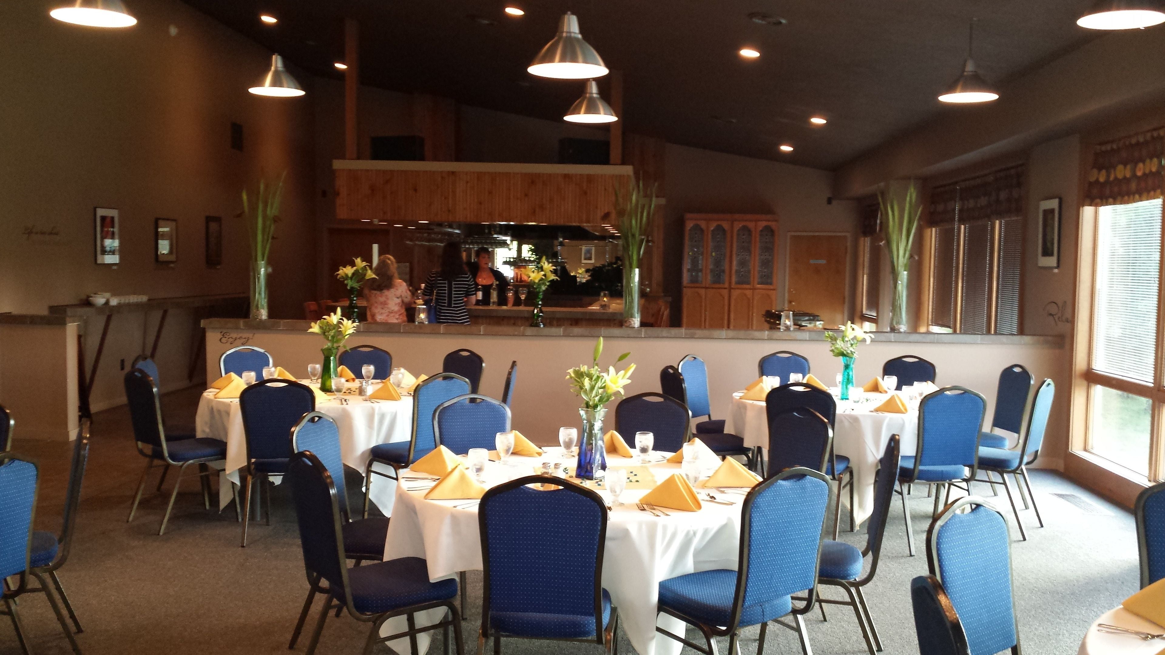 A dining area with round tables set with blue chairs and yellow napkins, featuring a kitchen in the background.