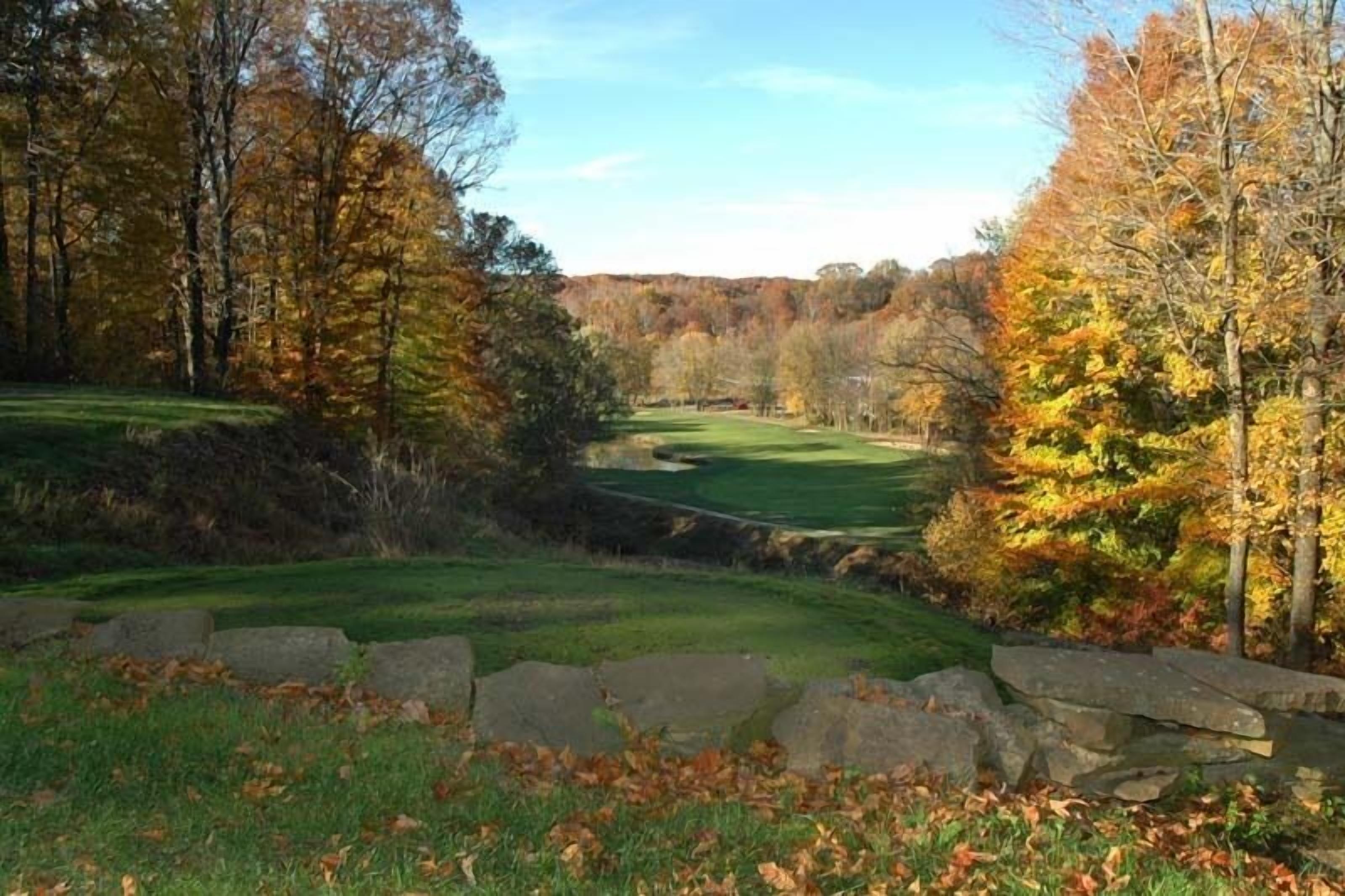 A golf course with a stone wall in the foreground, surrounded by trees with autumn foliage.