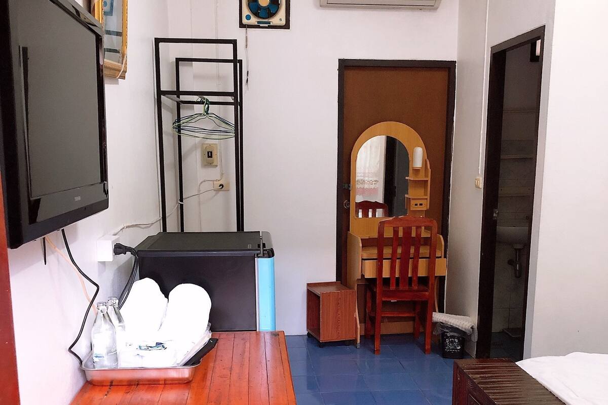 A room with a bed, a wooden table with a chair, a TV, an air conditioner, and a small mirror.