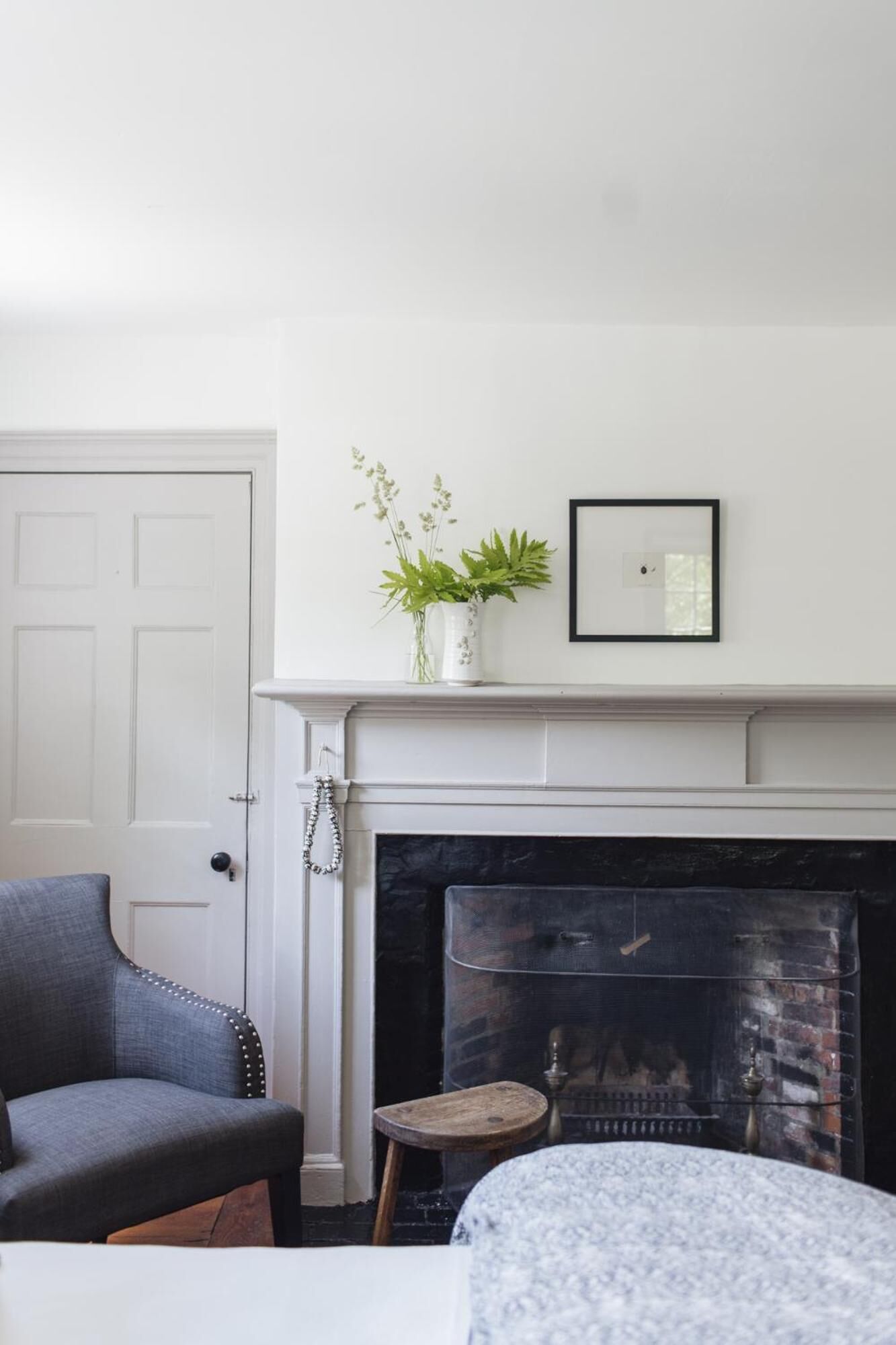 A room with a white door, a gray armchair, a small stool, a white fireplace mantel, and a framed picture above the mantel.