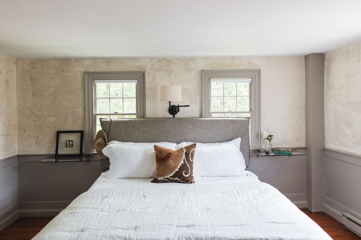 A bedroom with a large bed, two windows, a nightstand with a lamp, and a framed picture.
