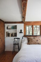Un dormitorio con techo de madera, una cama, una silla, un estante con libros y una ventana.