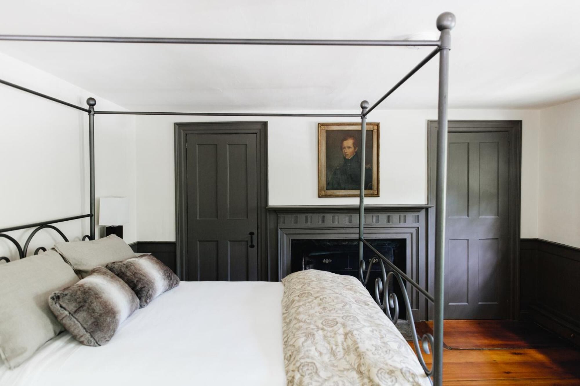 A four-poster bed with a white and patterned bedspread, two pillows, and a fireplace with a framed portrait above it.