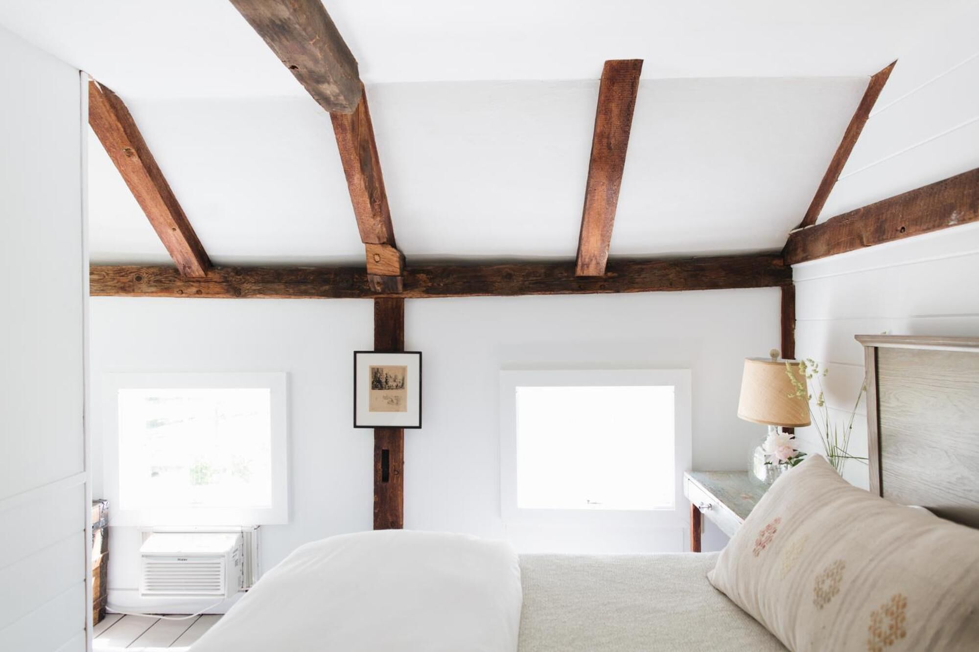 Un dormitorio con vigas de madera a la vista, una cama, una mesita de noche con lámpara y dos ventanas.