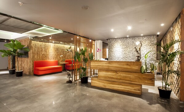 A modern reception area with a wooden reception desk, red seating, and potted plants. - Daun Bali Seminyak Hotel (Keroboken Kelod)