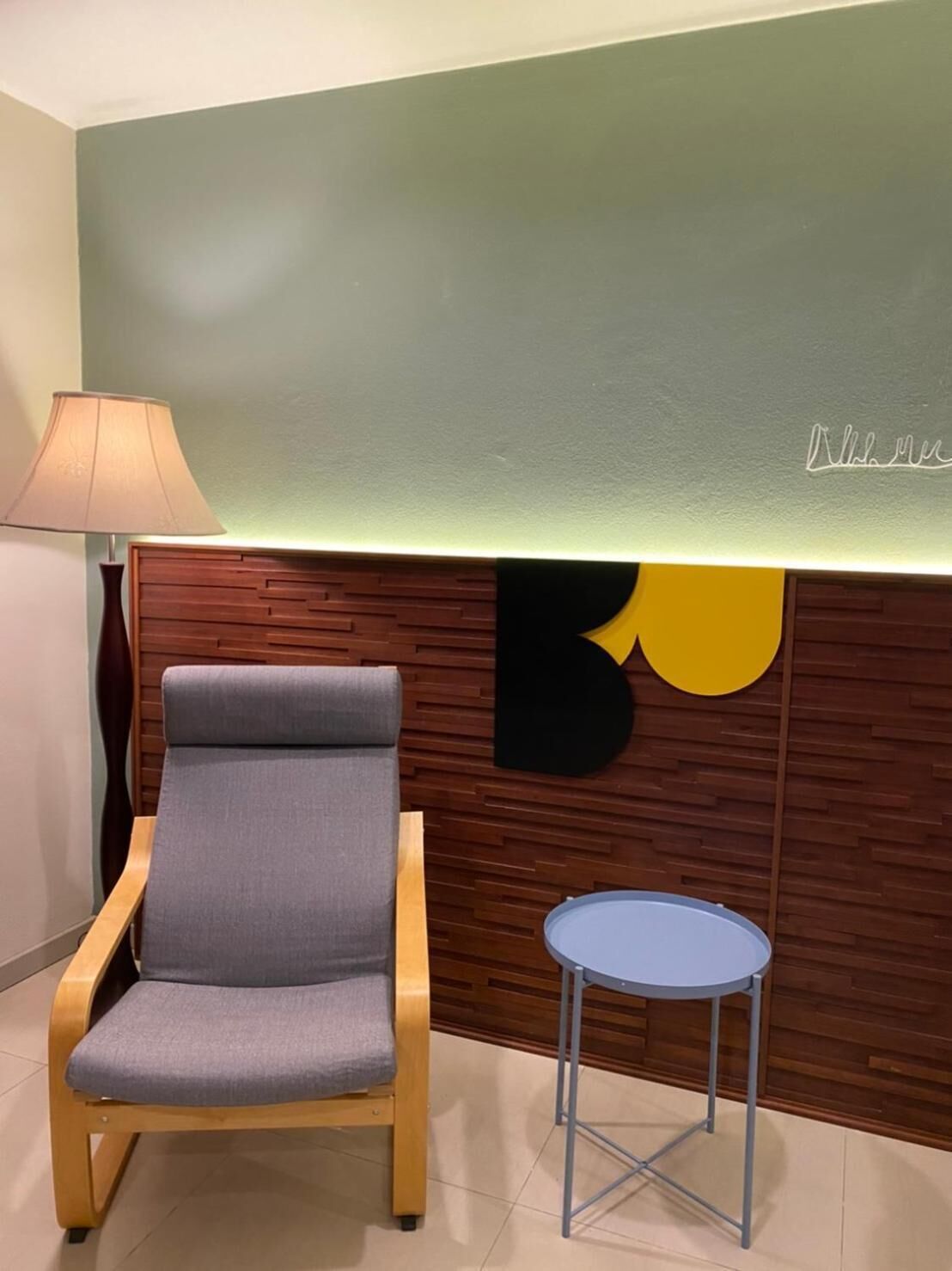 Una recepción moderna con un panel de madera, un sillón tapizado en gris, una mesita redonda y una lámpara.