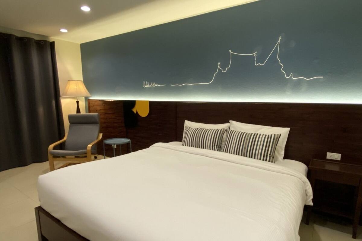 Une chambre d’hôtel avec un grand lit, une chaise, une petite table, une lampe et un mur orné d’un dessin linéaire.