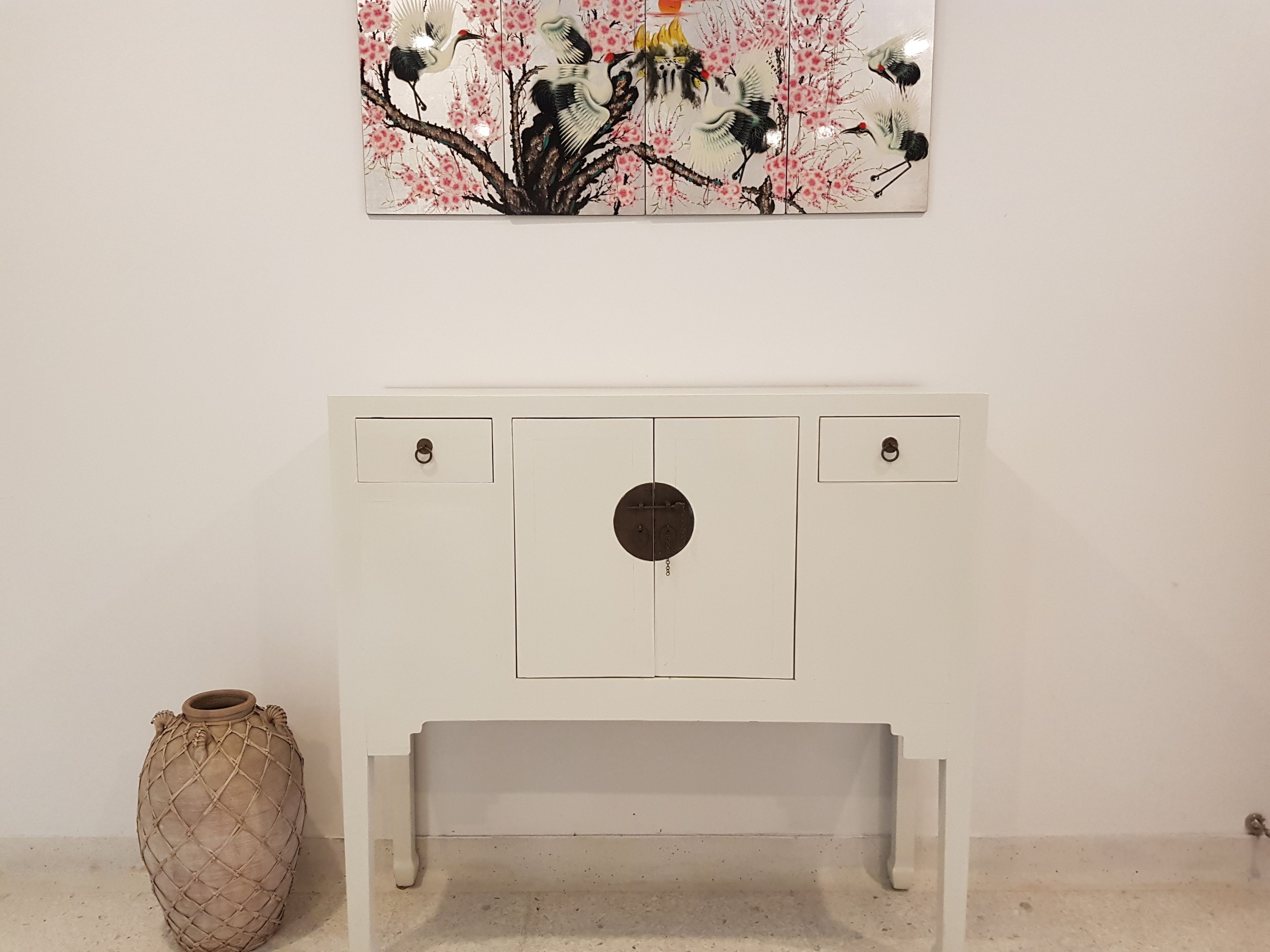 A white cabinet with a circular center panel and two drawers, a vase, and a painting of birds and cherry blossoms on the wall.