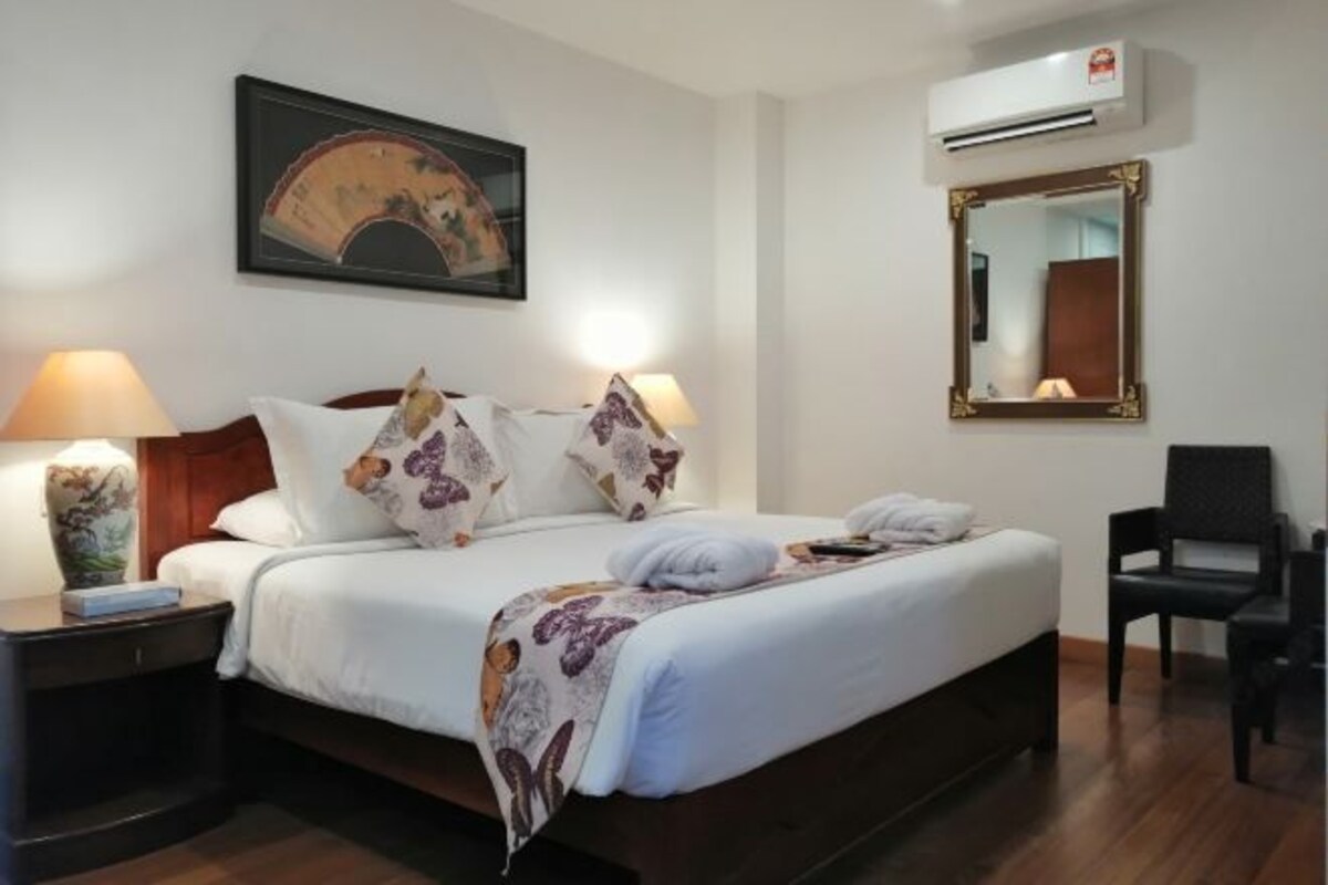 Een net opgemaakt bed met witte lakens en decoratieve kussens, een nachtkastje met een lamp, een wandairconditioner en een inlijsting spiegel.