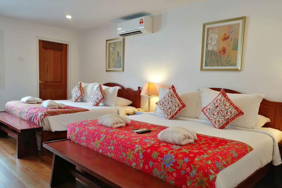 Une chambre d’hôtel avec deux lits, chacun recouvert d’une courtepointe à motifs floraux rouges, agrémenté d’oreillers blancs et doté d’une tête de lit en bois. Il y a un banc au pied de chaque lit, une table de chevet avec une lampe et un appareil de climatisation fixé au mur.