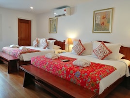 Habitación de hotel con dos camas, cada una con una colcha floral roja, almohadas blancas y cabeceros de madera. Al pie de cada cama hay un banco, una mesita de noche con lámpara y un equipo de aire acondicionado en la pared.