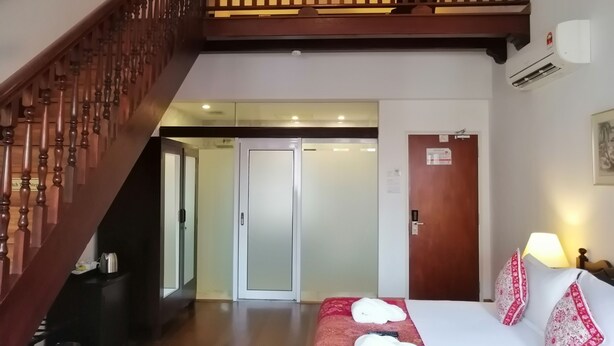Habitación de hotel con escalera de madera, cama con ropa blanca y almohadas rojas estampadas, mesita de noche con lámpara y puerta que da a un pasillo.