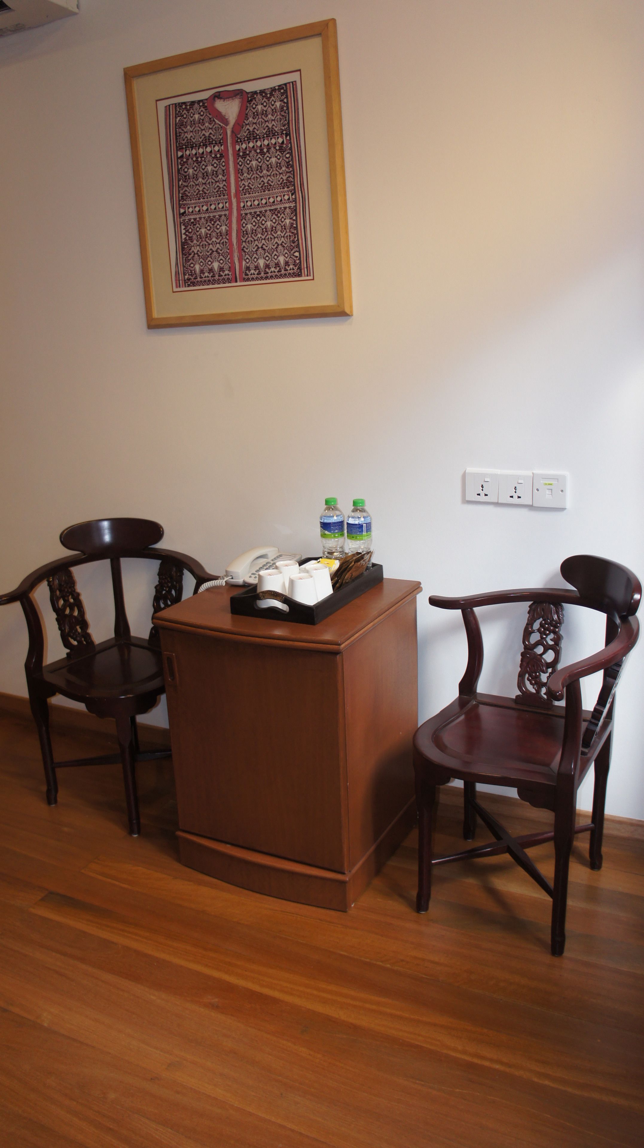 A room with two wooden chairs, a small wooden desk with a telephone and a tray of bottled water, and a framed artwork on the wall.