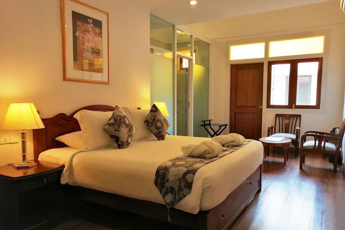 Une chambre d’hôtel avec un lit, une lampe de chevet, un plancher en bois, une chaise, une petite table et une fenêtre avec des stores.
