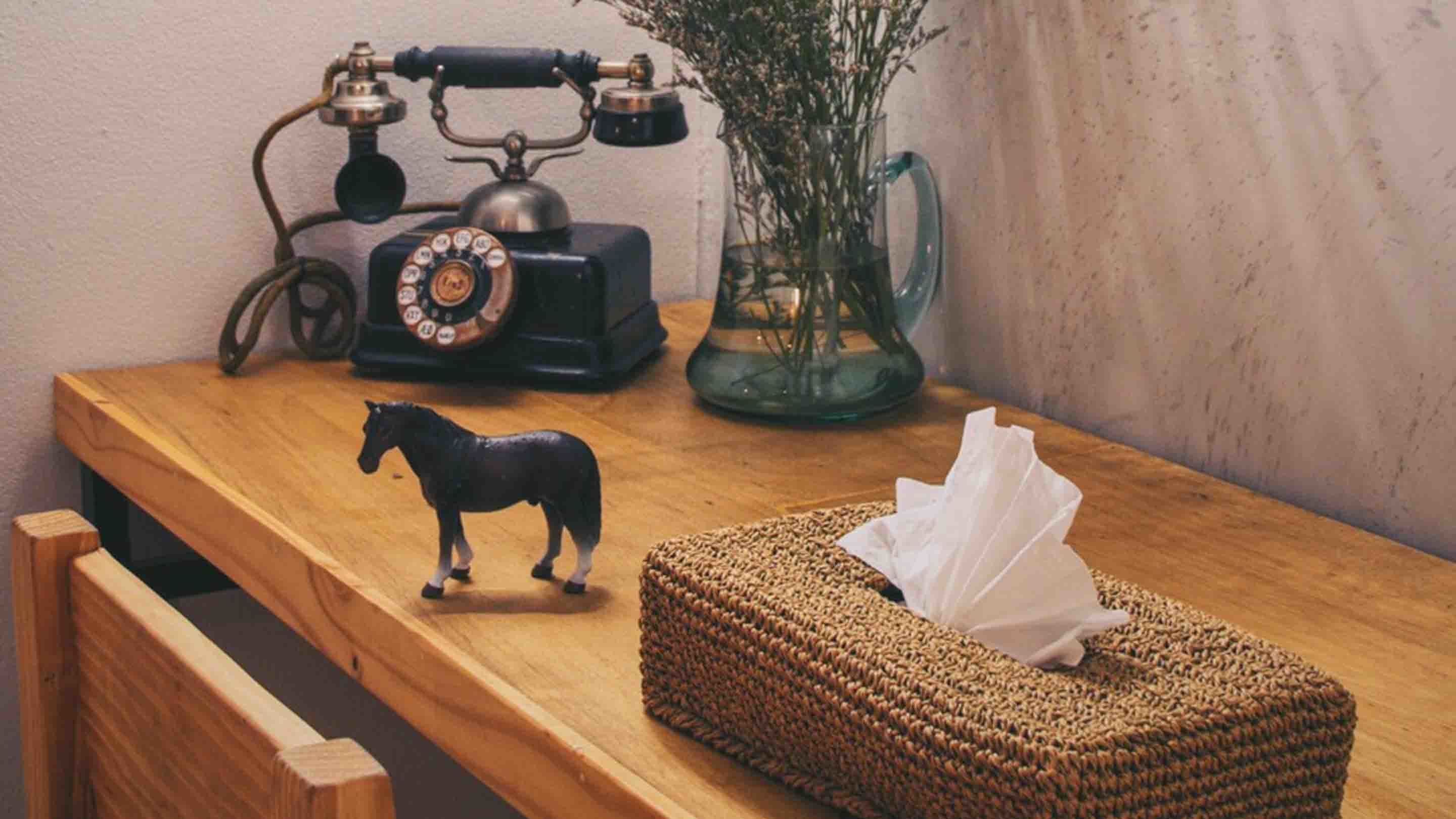 A wooden table with a vintage rotary phone, a small horse figurine, a woven basket, and a vase with dried flowers.