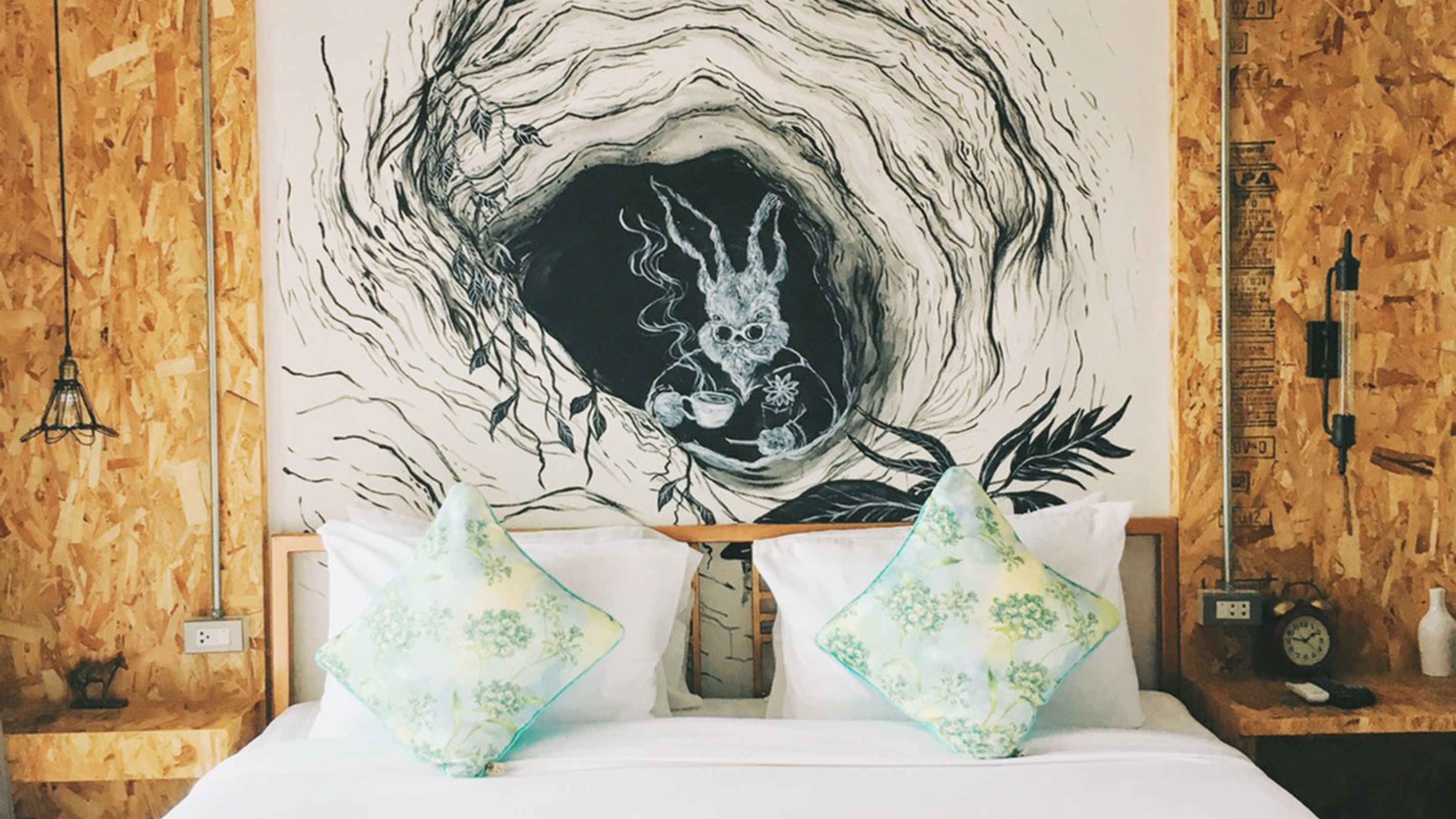 Un dormitorio con un amplio mural en la pared, paneles de madera, una cama con ropa de cama blanca, dos almohadas decorativas, una mesita de noche con un reloj y una pequeña estantería con una figurita.