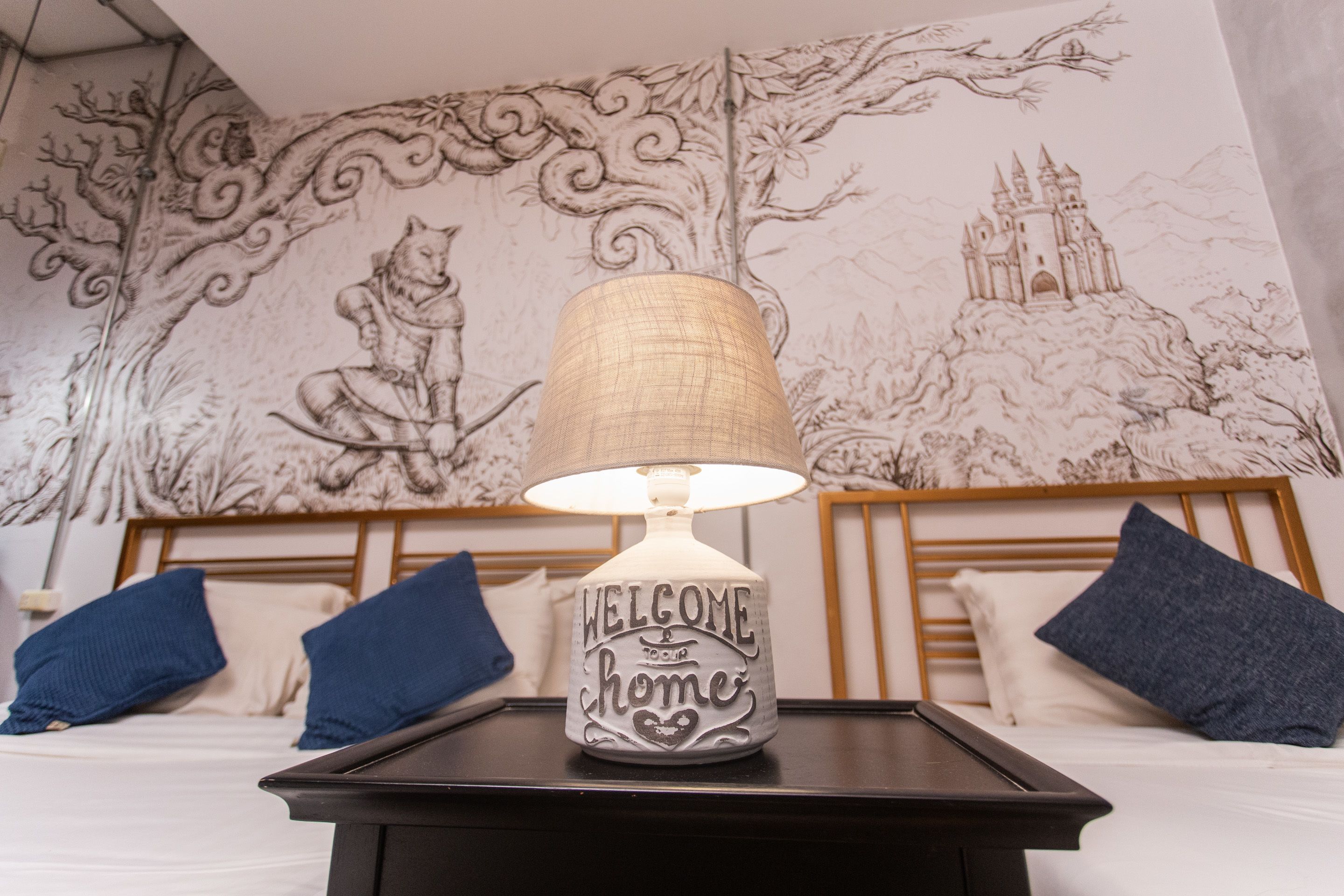 A lamp with "WELCOME HOME" on a bedside table in a room with a mural on the wall.