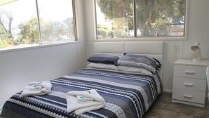 Una cama bien tendida con una colcha estampada, una mesita de noche con una lámpara y una ventana que da a árboles y edificios.