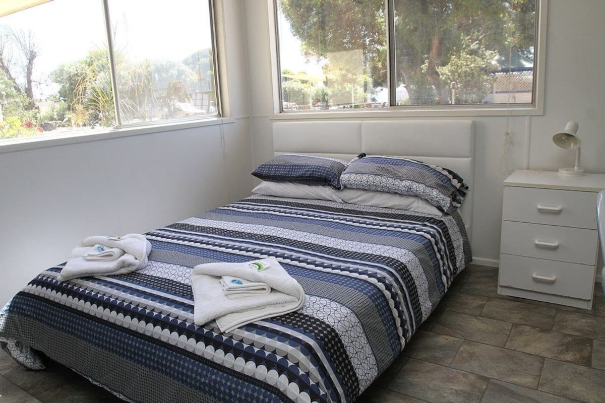 A neatly made bed with a patterned bedspread, a bedside table with a lamp, and a window with a view of trees and buildings.
