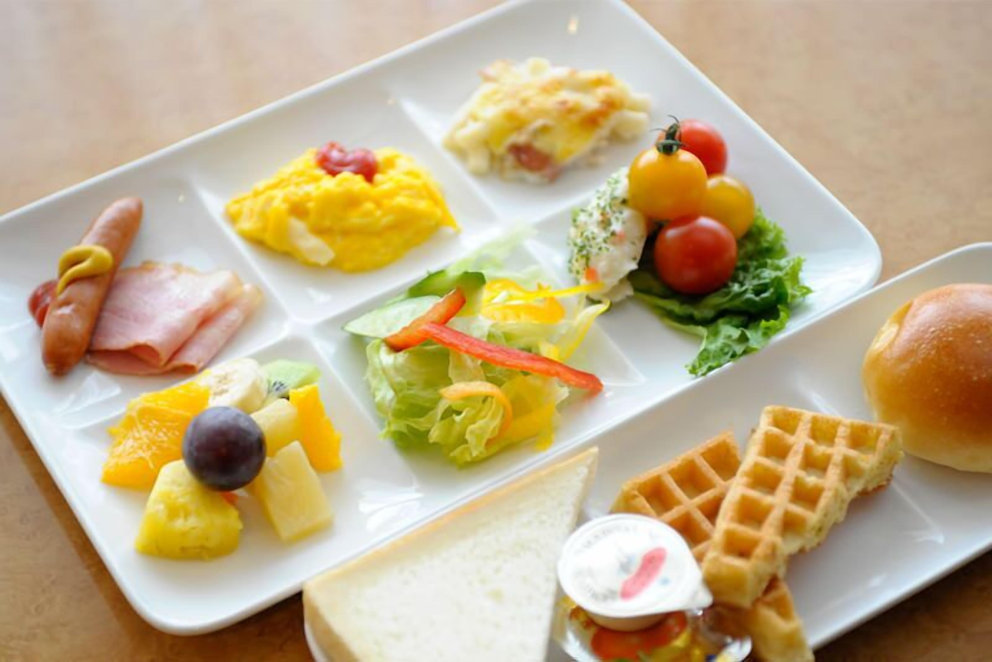 Un plato de desayuno con huevos revueltos, jamón, tocino, tostadas, gofres, fruta y una ensalada.