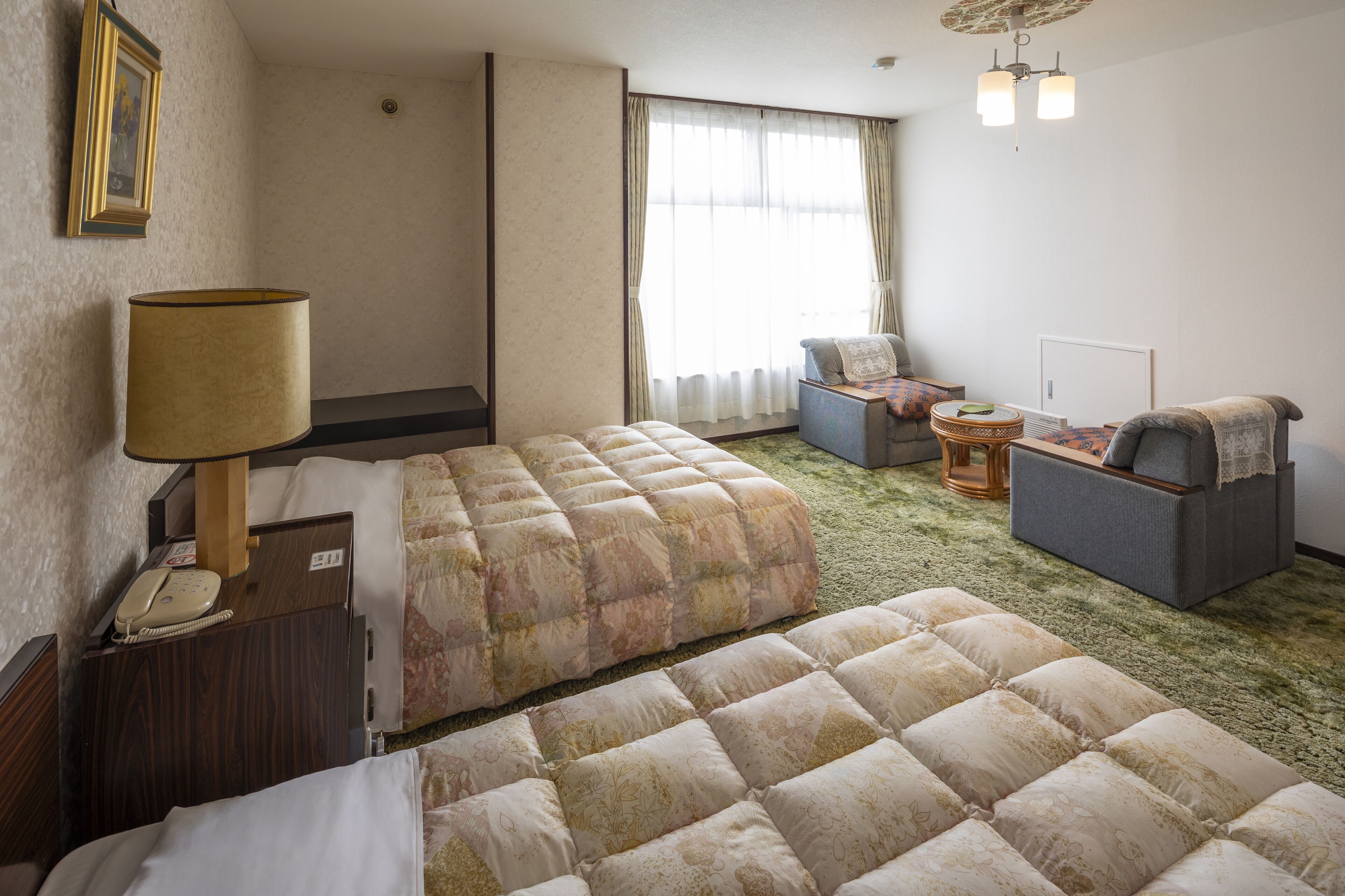 A hotel room with two beds, a desk with a lamp, a chair, a small table, and a window with curtains.