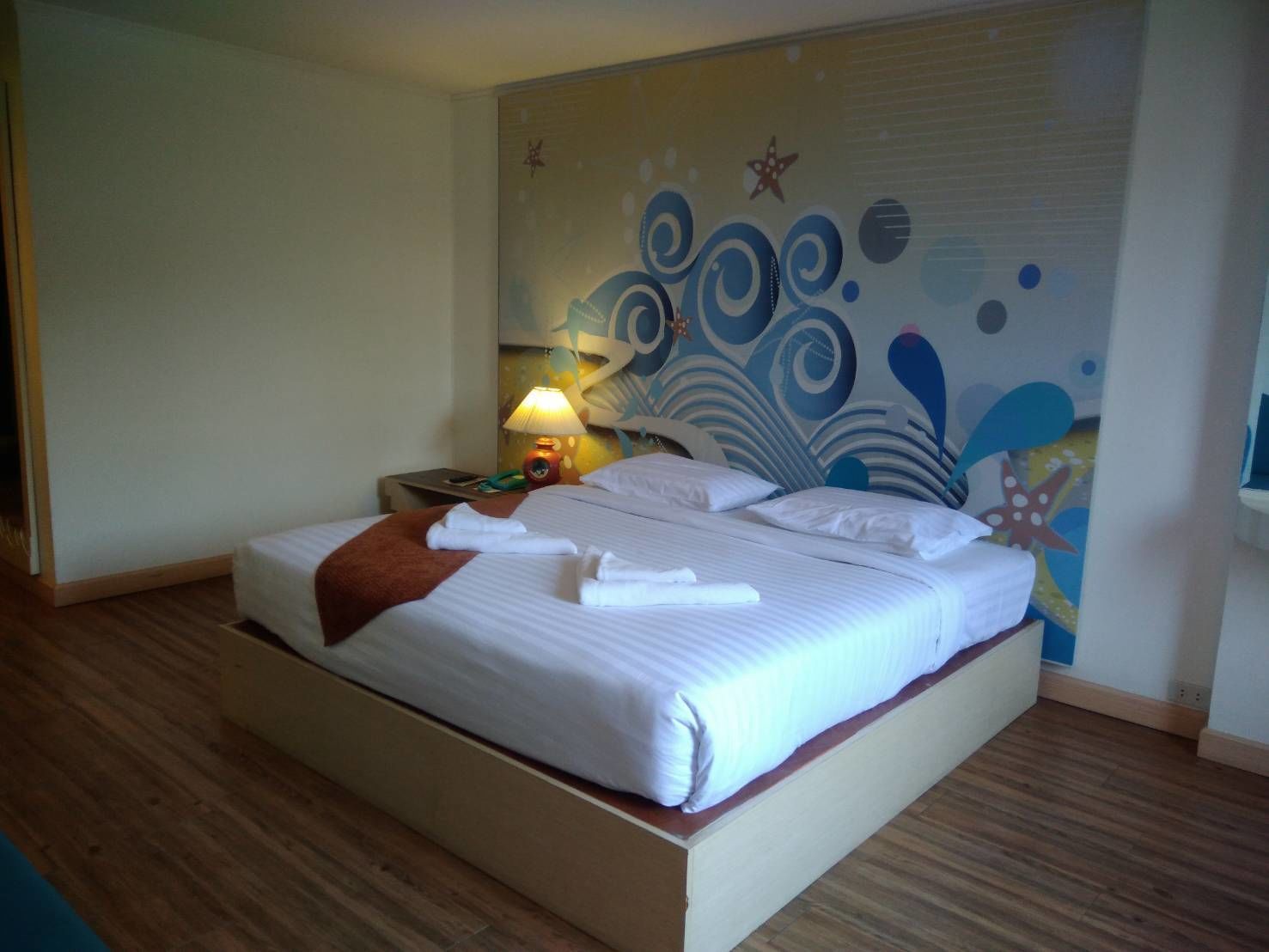Ein ordentlich bezogenes Bett mit weißen Laken und einer braunen Tagesdecke, eine Nachttischlampe und eine Wand mit einem Seestern und einem abstrakten Design.