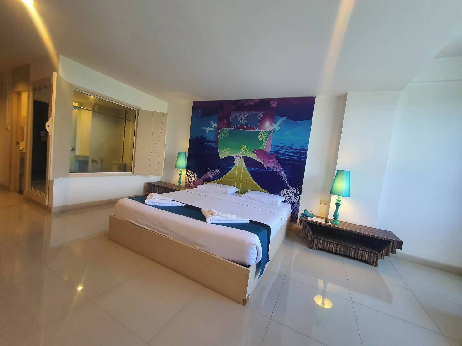 Habitación de hotel con una cama grande, dos mesitas de noche con lámparas, un espejo amplio y un mural colorido en la pared.