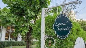A sign for 'Lovin' Sweets' with a decorative metal frame and a bell hanging below it, set against a backdrop of lush greenery and a building with large windows. - The Pacific Cool Chic (Chiang Mai)