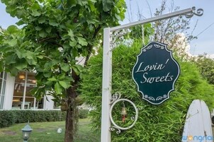 A sign for 'Lovin' Sweets' with a decorative metal frame and a bell hanging below it, set against a backdrop of lush greenery and a building with large windows.