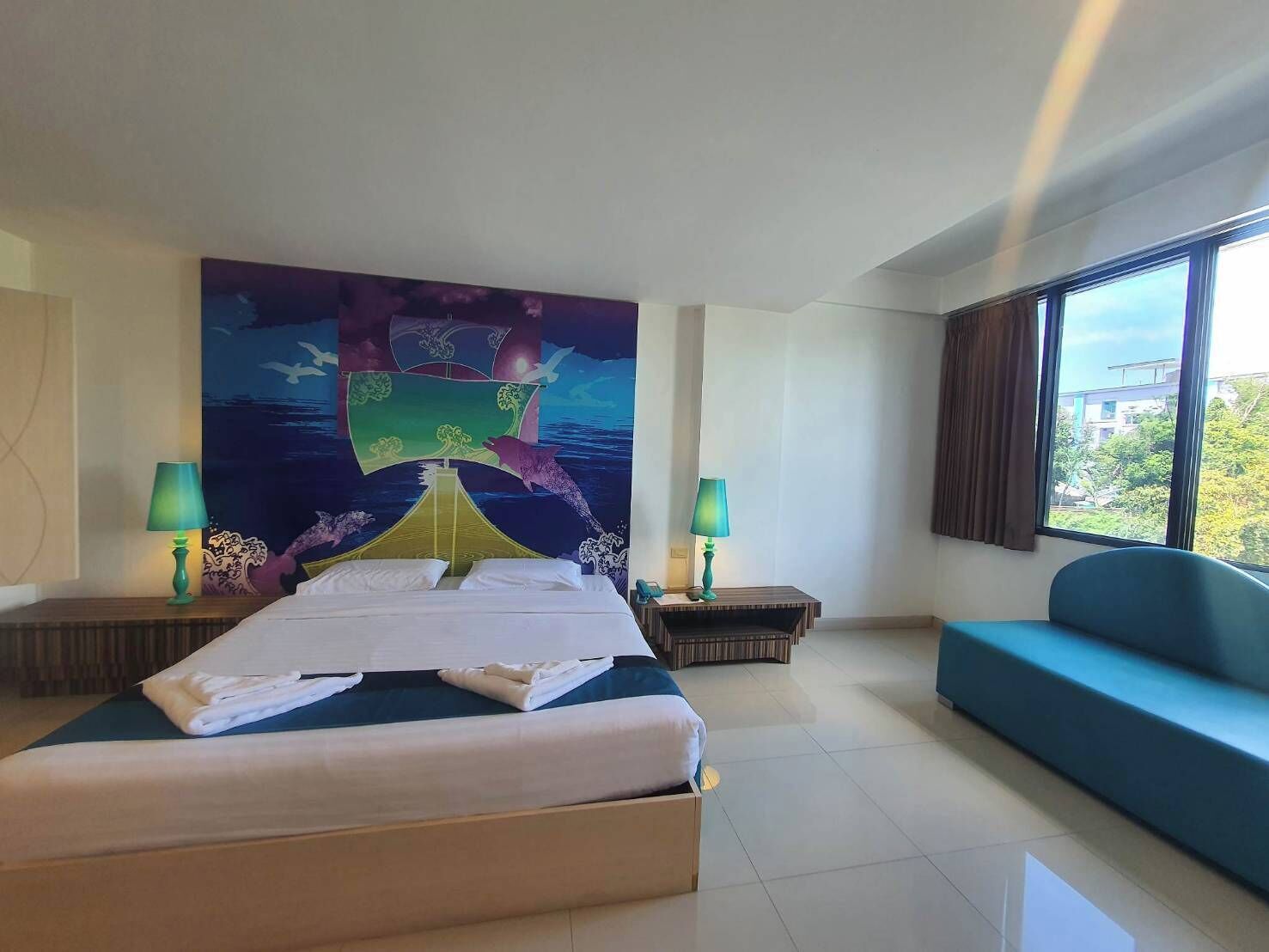 A hotel room with a bed, two bedside tables with lamps, a sofa, and a large mural on the wall.