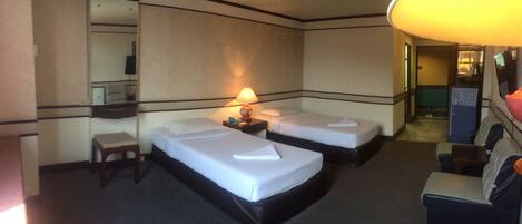 A hotel room with two beds, a desk, a chair, a lamp, and a sofa.