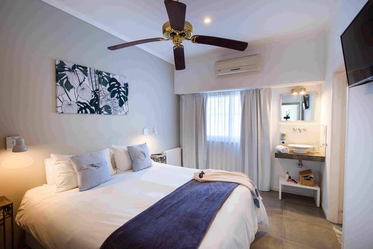 A bedroom with a bed, a ceiling fan, a painting, a bathroom with a sink and mirror, and a window with curtains.