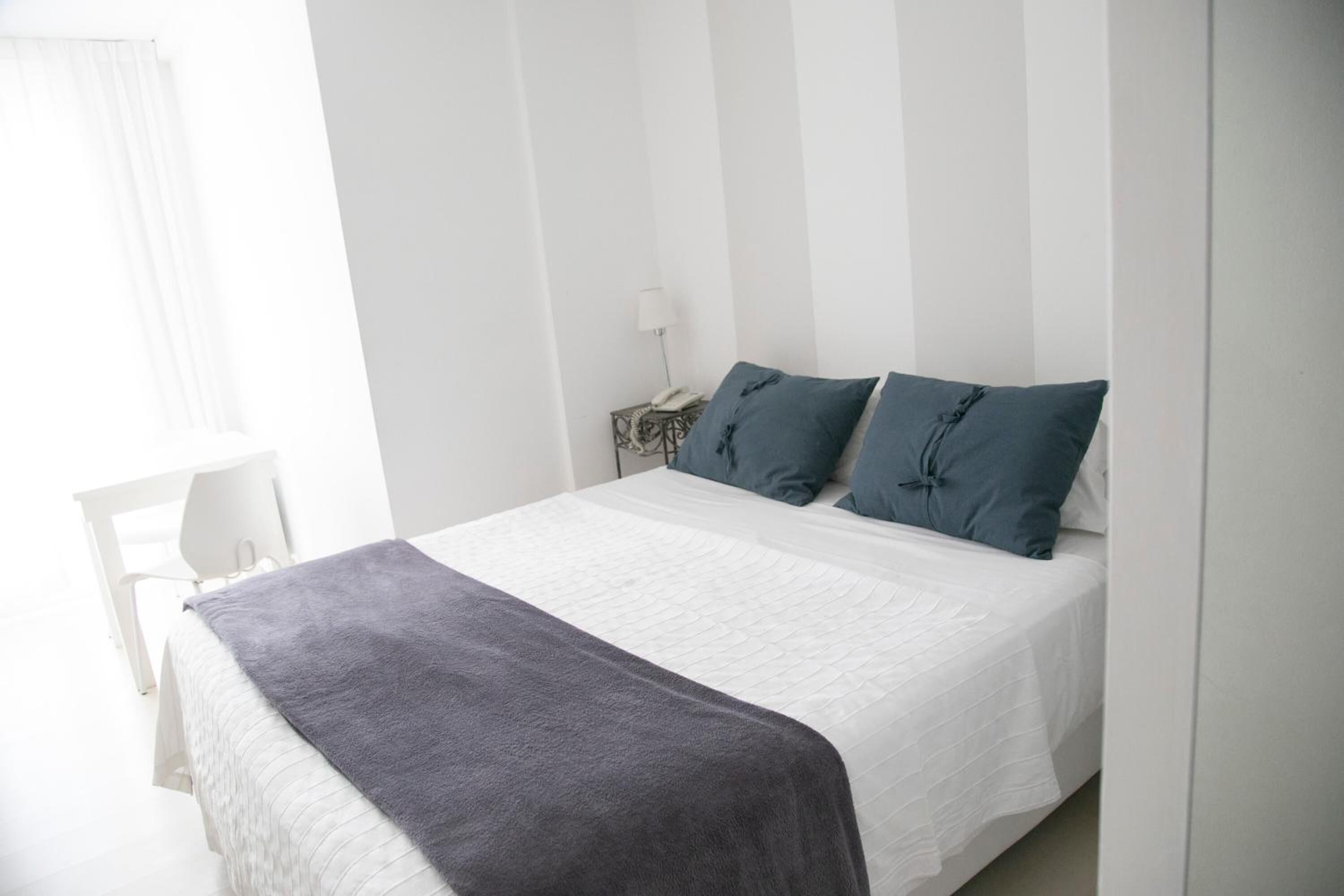 Un lit bien fait, avec une couette blanche et un plaid gris, deux oreillers bleus et une table de chevet blanche sur laquelle se trouve une lampe.