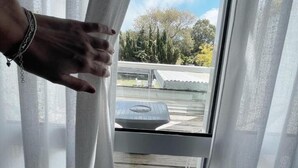A hand adjusting a window blind in a room with a view of a wooden deck and trees outside. - Hotel Valeria (Valeria del Mar)