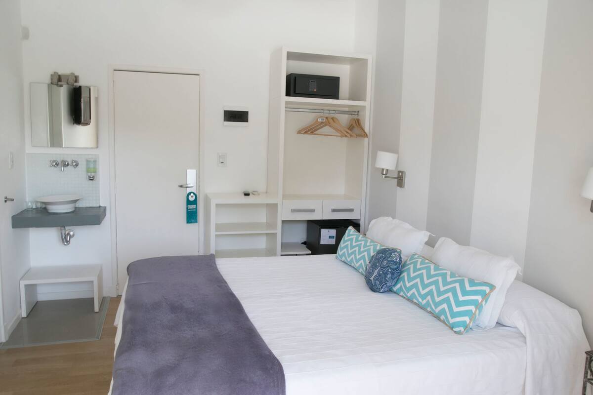 A bedroom with a bed, a bedside table, a mirror, a wardrobe, and a bathroom with a sink and towel rack.