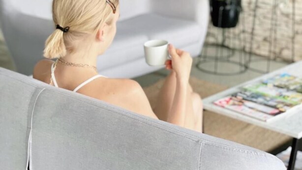 A person sitting on a grey sofa, holding a white mug, with a magazine on a table in front of them. There are two potted plants on a wall behind the sofa. - Hotel Valeria (Valeria del Mar)